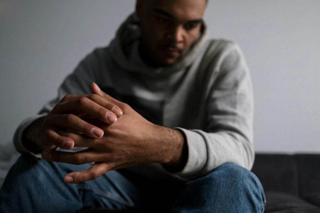 A man in a gray hoodie sits with his hands clasped together in the foreground, head bowed and face blurred in the background, conveying a mood of worry, sadness, or deep thought.
