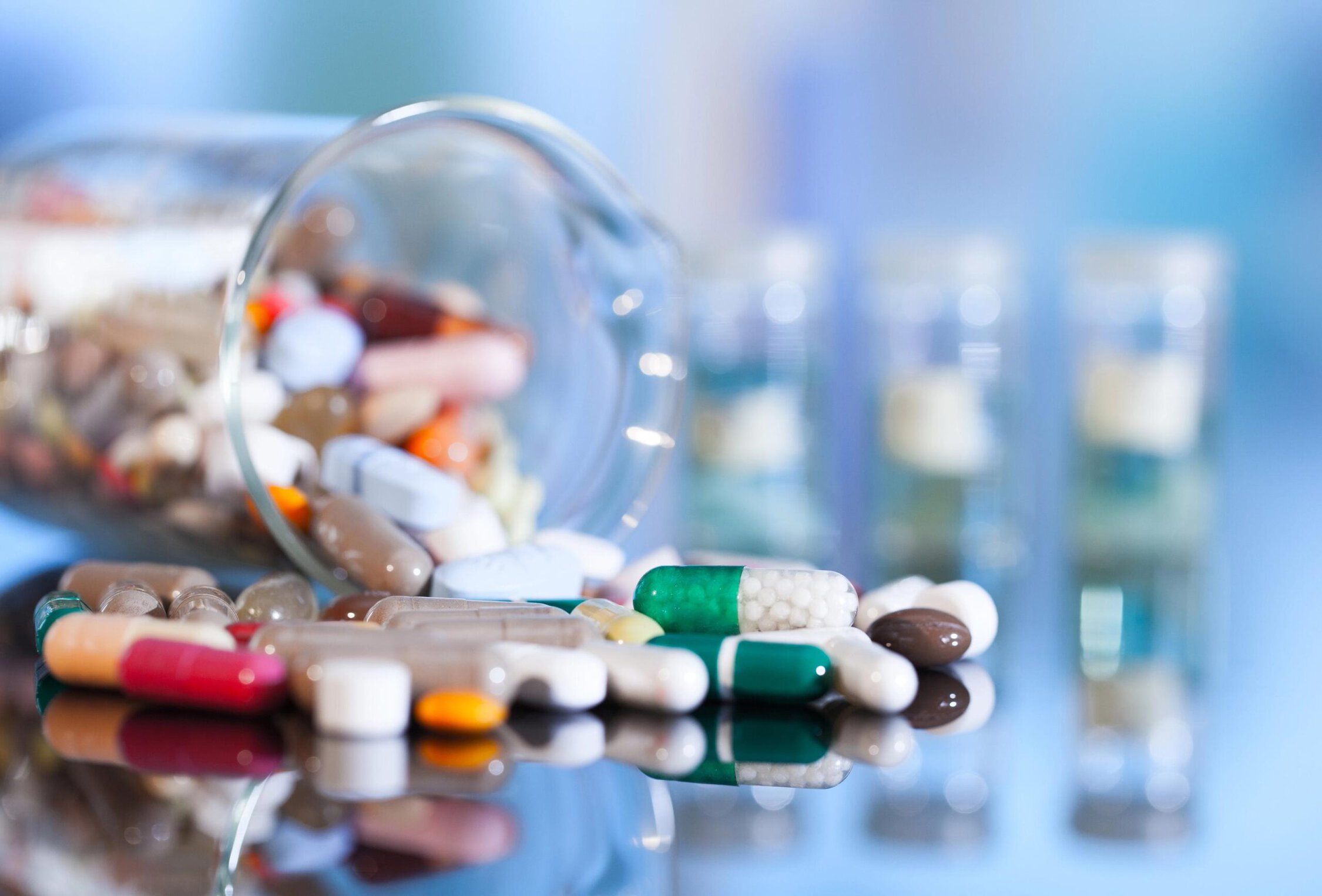 Assorted pills spilled from a glass container on a reflective surface.