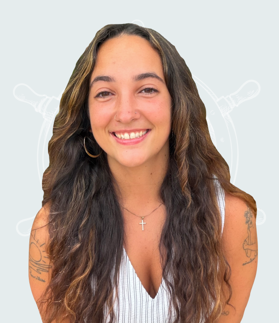 Smiling young woman with long wavy hair and tattoos on arms in front of a nautical background.