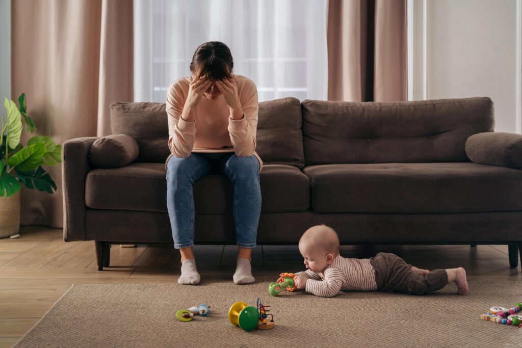 Woman distressed on sofa with baby playing on rug in living room.