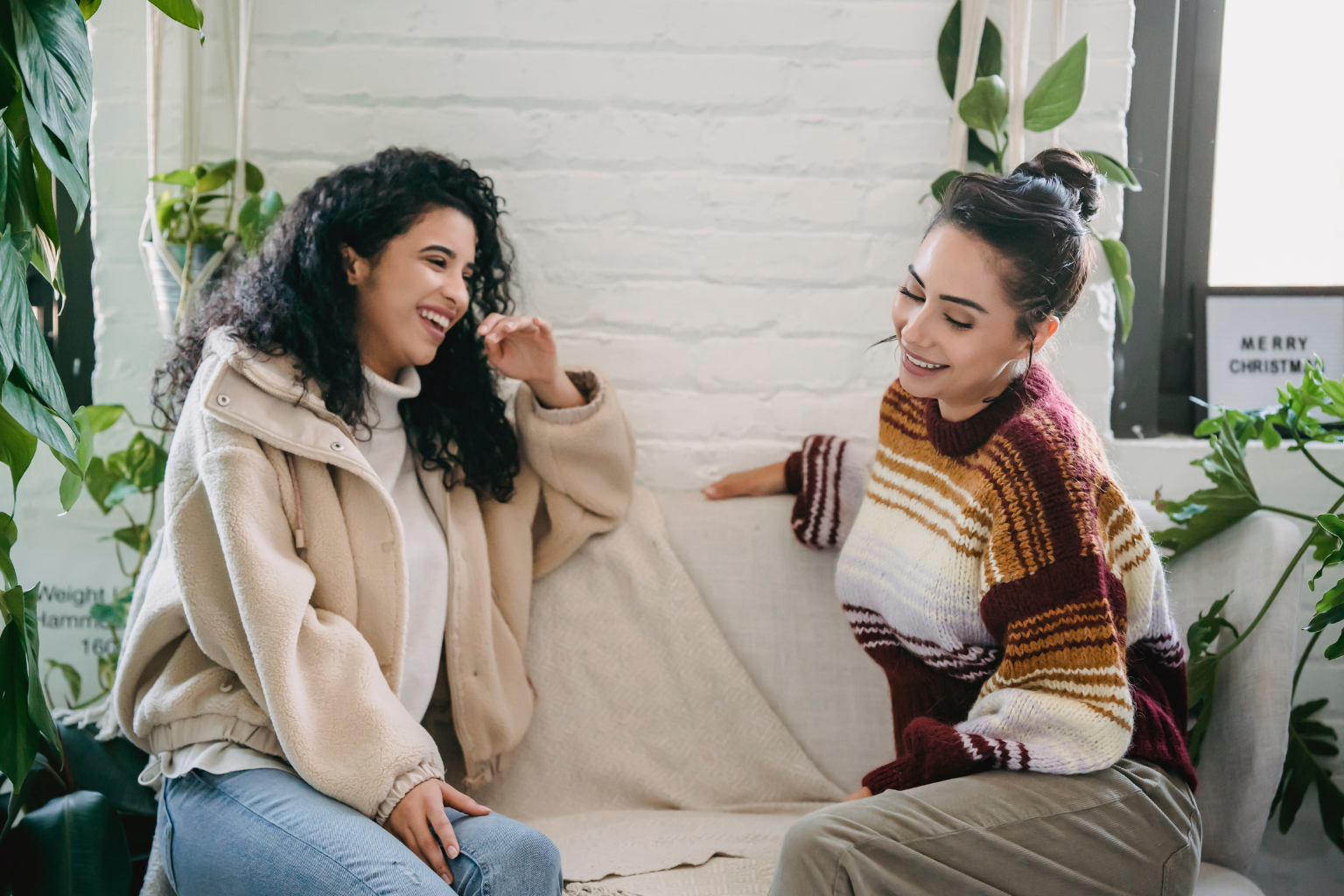 Two women smiling and talking in a cozy, plant-filled room.